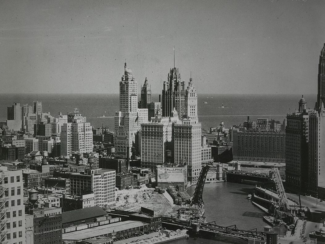 A wide, high-angle black-and-white photograph of the Chicago skyline and riverfront. The image shows historic skyscrapers, including the Tribune Tower, with industrial areas and drawbridges in the foreground 