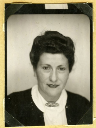 A black and white studio portrait of a young Rodi, likely from the mid-20th century. She has short, dark, wavy hair styled with a small curl pinned at the top. She is wearing a dark plaid dress with a white Peter Pan collar. She looks directly at the camera with a soft, slight smile and dark, expressive eyes. The background is a solid dark color.