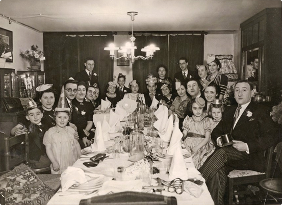 A wide black-and-white photograph of Rodi's large, multi-generational family seated at a long formal dinner table. Many of the guests, including the children and adults, are wearing festive paper party hats. The table is set with white linens, tall napkins, and glass bottles. A large chandelier hangs overhead, and the room is decorated with flowers and traditional wooden furniture. The atmosphere appears celebratory and crowded.