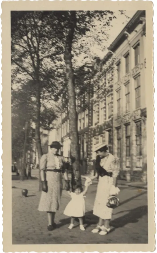 A vertical, vintage black-and-white photograph with a scalloped white border. Rodi's mother and aunt dressed in fashionable mid-length patterned dresses, gloves, and hats, walk along a brick-paved sidewalk. They are holding the hands of a young Rodi who is wearing a white dress and is stepping forward between them. Tall, narrow European-style buildings and leafy trees line the background of the street.