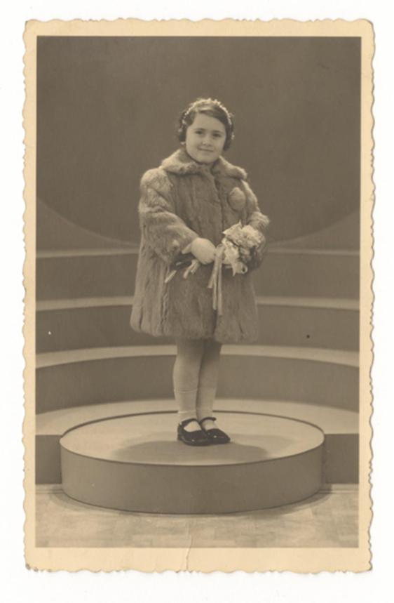 A studio portrait of a young  Rodi, standing on a low, circular stage in front of a background with curved, stepped lines. She is wearing a thick, textured fur coat, white socks, and dark Mary Jane-style shoes. She holds a small bouquet of flowers in front of her and has a decorative headband or floral crown in her hair. 