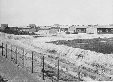A wide-angle black-and-white photograph of Westerbork transit camp, showing a series of long, low wooden barracks behind a barbed-wire fence