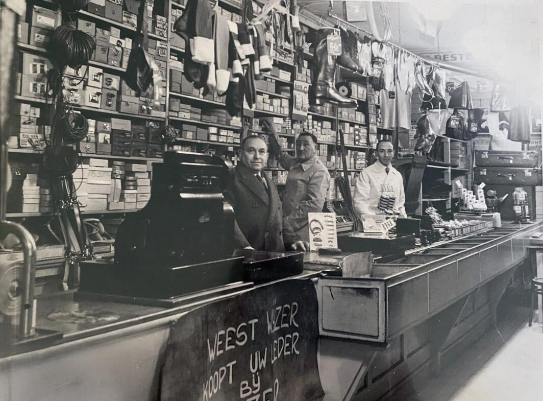 A black-and-white photograph of the interior of the Waterman family store. A long, dark wooden counter runs across the foreground, featuring a vintage mechanical cash register. Three men are positioned behind the counter; one is in a suit, while another wears a white work coat. The walls behind them are floor-to-ceiling shelves packed with boxes. Various leather goods, including boots, belts, and suitcases, hang from the ceiling and are stacked on shelves. A sign in Dutch on the front of the counter reads 'WEEST WIJZER KOOPT UW LEDER BIJ...'.