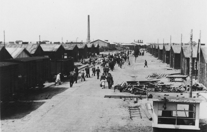 A historical photograph of the central road at Westerbork transit camp, showing people walking between rows of barracks and train cars.