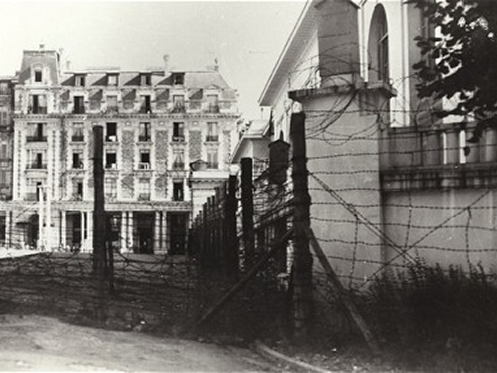 A black-and-white photograph showing the perimeter of the Vittel internment camp in France during WWII. In the foreground, several rows of tall barbed-wire fencing and wooden posts create a barrier across a dirt path. In the background, the ornate, multi-story architecture of a grand hotel stands in sharp contrast to the prison-like fortifications in front of it.