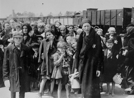 A black-and-white historical photo of a crowd of people, including women and children wearing Star of David badges, standing near wooden train cars.