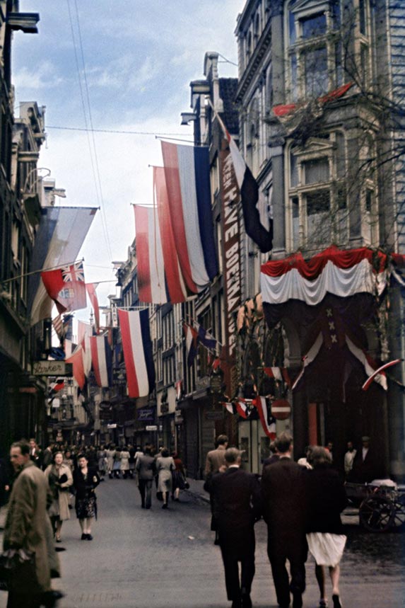 A vibrant colorized photo of Kalverstraat in Amsterdam on June 27, 1945. The narrow, historic street is crowded with people walking and is festively decorated with numerous large Dutch national flags (red, white, and blue) hanging from the buildings. An Allied flag, possibly British, is also visible. The atmosphere appears celebratory following the end of the war, with people dressed in mid-1940s attire.