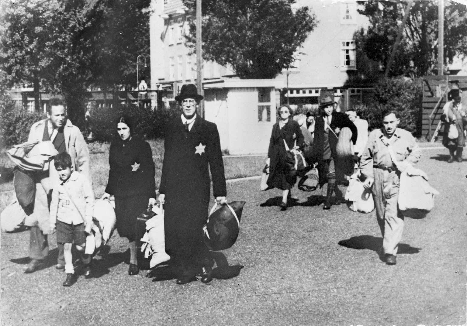 A historical photograph from June 1943 in Amsterdam. A group of Jewish men, women, and a small child walk toward an assembly point at Olympiaplein square. They are carrying heavy suitcases and bundles of belongings. Most of the adults have a visible yellow Star of David sewn onto their dark coats. The expressions are somber as they are forced to leave their homes under Nazi occupation.