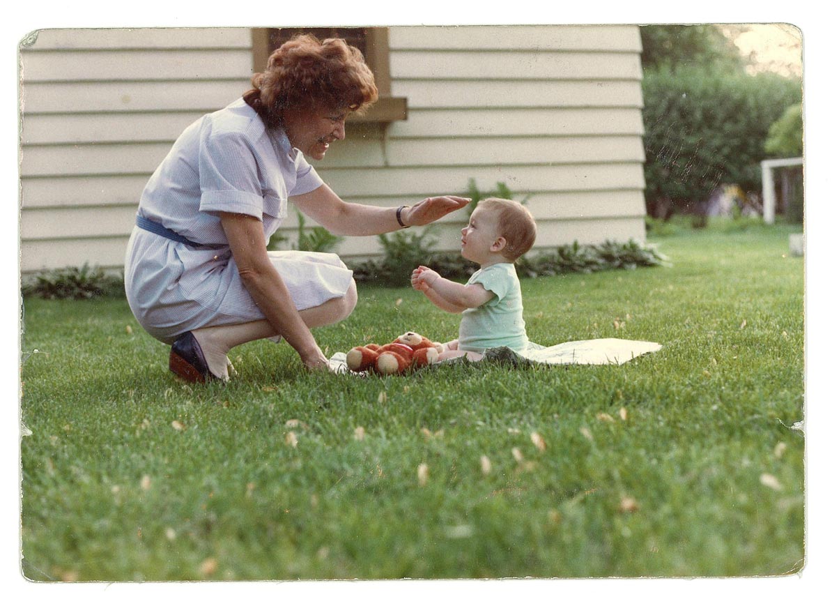 A color photograph of Fritzie in a light-blue striped dress kneeling on a green lawn, playfully interacting with her grandson who is sitting on a white cloth with a stuffed bear. 
