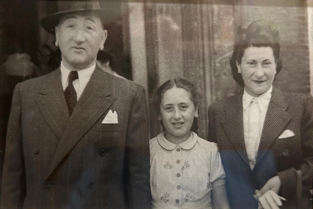 A black-and-white postwar photograph of Rodi and her parents. On the left, Rodi's father wears a dark suit, tie, and a fedora. In the center, Rodi, smiles gently, wearing a light-colored, short-sleeved blouse with a floral pattern and a Peter Pan collar. On the right, Rodi's mother wears a dark patterned blazer and a light blouse, her hair styled in a typical 1940s fashion.