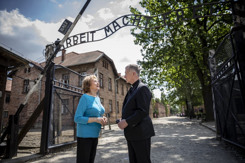 A modern color photo of Fritzie in a blue shirt speaking with a Cardinal Cupich, who is wearing a black clerical suit, as they stand under the 'Arbeit Macht Frei' gate at the Auschwitz memorial site. 